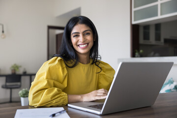 Indian woman sits at table with laptop engaging in professional tasks, working in homeoffice, feel happy and satisfied with productive workflow, take break, look at camera, posing. Freelancer portrait