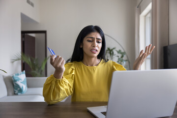 Indian woman looks at laptop screen with angry expression while holds card, try to make payment unsuccessfully, feels concerned due to additional fees or shipping costs during checkout, card limit