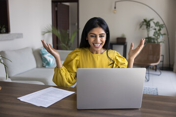 Overjoyed Indian woman sitting at work desk looking at laptop screen with amazed, surprised...