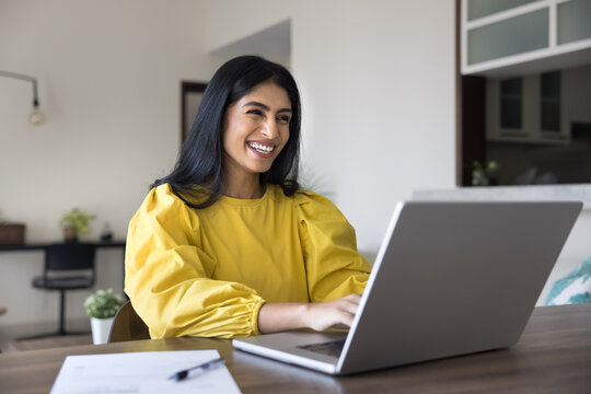 Happy Indian woman sit at table with laptop, working remotely, engage in professional tasks, browsing information using modern device and internet connection, taking online course, respond to messages