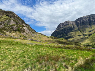 View of mountain landscape in Glencoe, Scotland. Green hills and a scenic valley under a blue sky with clouds.