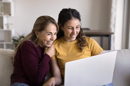 Two different generations women looking at laptop screen seated on sofa, review new AI platform, shopping online, browsing products, discussing options, or making purchases, video-calling to family