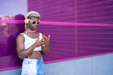 Stylish non-binary person wearing headphones and using smartphone in front of purple wall