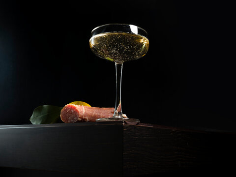 Low angle close-up of a glass of champagne in a champagne coupe and salami sausage on a table with a lemon