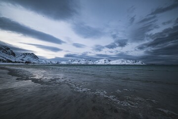 Serene winter seascape with snow-covered mountains under a dramatic cloudy sky in Lofoten island