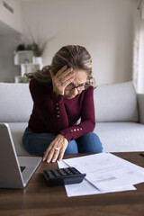 Sad woman sits on couch, hold her forehead in distress while looks at financial documents spread...