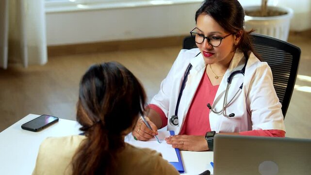 Indian Asian young female doctor writing a medical case report, diagnosis, prescription for a senior woman patient sitting in lavish clinic, carefully documenting health records for accurate treatment