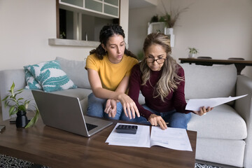 Latina mother and her grown up daughter managing family finances together sit at table with laptop, calculator and heap of household utility bills to pay, review incomes and expenses, planning budget