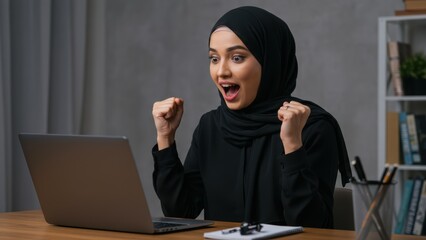 Young Arab woman in hijab cheering at laptop in home office Celebrating success and engagement in digital workspace