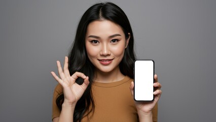 Fototapeta premium Asian woman holding smartphone, showing okay gesture, with neutral background, representing modern lifestyle and digital communication