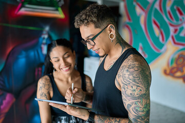 Latin tattoo artist showing tattoo design to smiling client on digital tablet in colorful studio