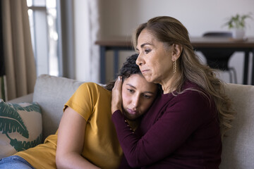 Loving aged mother hugging, comforting her young adult daughter seated together sofa, girl...