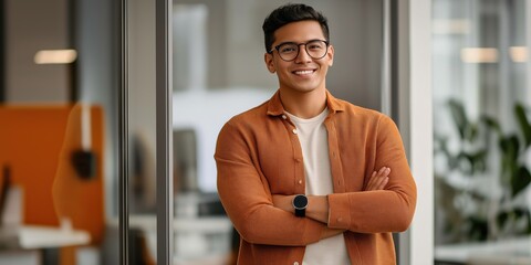 A man in an orange sweater and glasses is smiling and standing in front of a window. He is wearing a watch and has his arms crossed