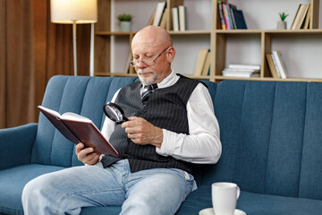 Mature man reading a book with magnifying glass on the sofa in living room