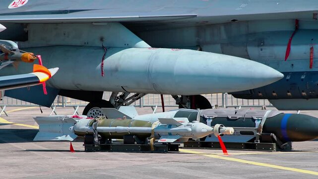 Detailed view of a military aircraft's underwing pylons with mounted fuel tanks, missiles, and bombs, displayed on ground with safety ribbons and securing straps