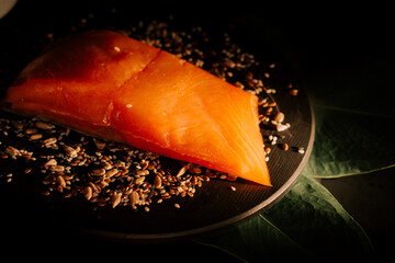 Fresh organic trout fillet resting on black board surrounded by various seeds and leaves in a dimly...