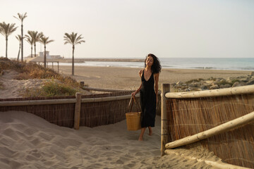 Brunette Woman in Black Dress Walking on Sand Dunes at the Beach with Straw Bag