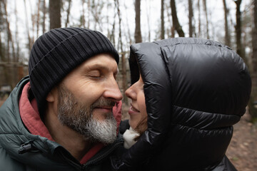 A couple stands close in a forest, sharing a joyful moment while wearing winter attire. The trees...