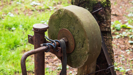 Rusty sharpening stone on a wooden pole in the woods. The sharpening stone is partially covered with moss and dirt. A metal rod with a handle, which was used for turning, comes off of it.