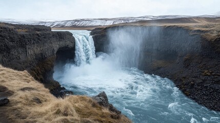 Obraz premium Majestic Icelandic Waterfall Cascading into a Turbulent River Surrounded by Dramatic Cliffs and Winter Scenery Under a Gloomy Sky