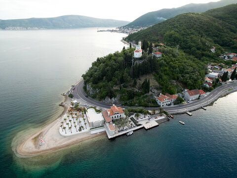 Embankment of a green cape with houses surrounded by forest, a lighthouse and a church on a hill. Lepetane, Montenegro