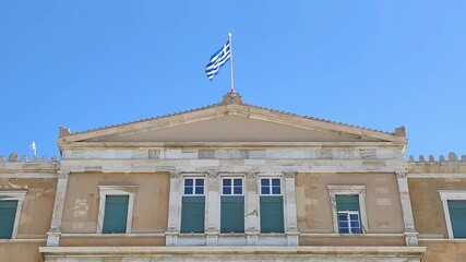 Video of the Greek flag waving atop the historic Greek Parliament building in Athens, Greece, symbolizing national pride and heritage against a clear blue sky. - Powered by Adobe