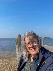Frau mit langen grauen Haaren sitzt am Steg am Meer, ihre Haare sind vom Wind zerzaust