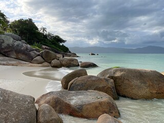 Big boulders in the late afternoon sun. Brown and orange stones, green turquoise sea, sky full of gray and white clouds, mountains in the horizon, green vegetation, sand. Jurere Internacional beach.