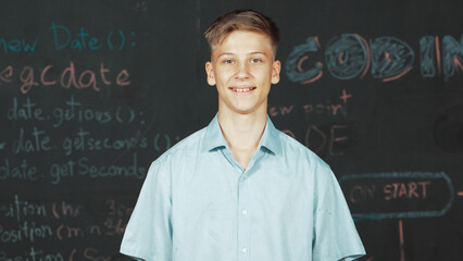 Smiling teenager looking at camera while standing at blackboard with engineering code or prompt...
