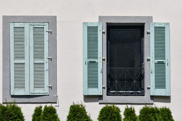 A pair of windows with stone frames and pale green shutters, one window closed and the other open, on the white facade of an elegant home, with the top of a hedge in the foreground, France