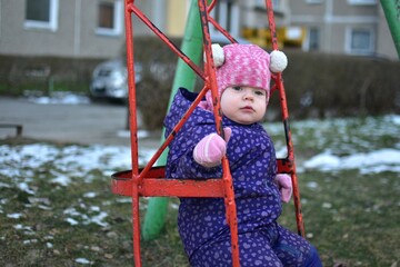 Baby girl swinging in soviet style old children playground in cold weather. Toddler girl having fun in public playground in winter