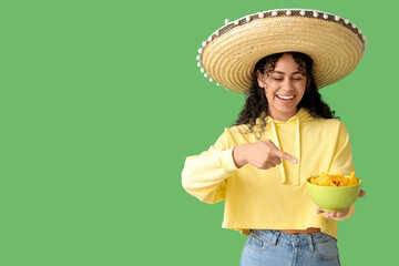 Beautiful young African-American woman in sombrero hat pointing at bowl with tasty nachos on green...