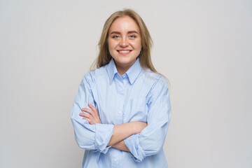 Smiling young woman poses confidently with arms crossed in a light blue shirt against a neutral...
