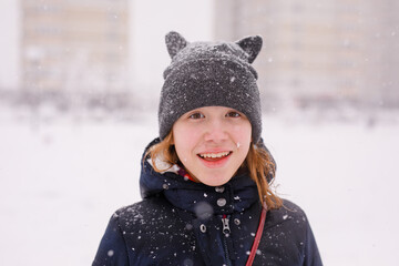 Portrait of an unhappy 12-year-old girl in a warm jacket and a cat-ear hat under heavy snow