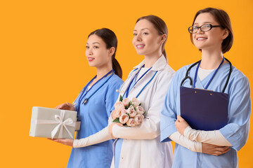 Obraz premium Female doctors with gift and tulips on yellow background. International Nurses Day