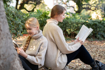 Woman and girl sitting back to back, using a tablet and reading a book in a park during a sunny autumn day, enjoying some quality time together