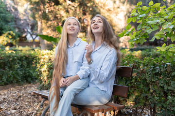 Mother and daughter with long blond hair, dressed in casual clothes, sitting together on a park...