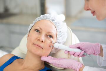 Young female cosmetologist performs facial microcurrent procedure with machine to adult female...