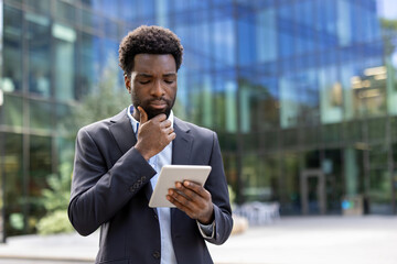 Serious thinking businessman with tablet computer in hands outside office building. African...