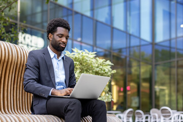 A young professional works remotely, utilizing his laptop while seated on a modern bench with an...