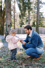 Smiling dad squatting near a little girl examines a fir cone in his hands in a spring park