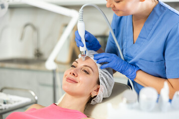 Adult woman cosmetologist performs hardware facial procedure to young woman patient