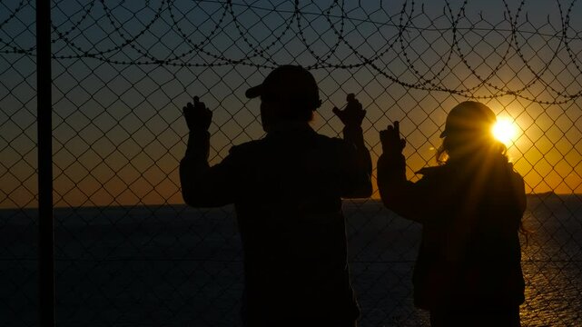 Refugees looking at sunset behind barbed wire. Refugees standing silently, silhouetted against warm sunset light, gazing through barbed wire fence with deep longing for freedom and hope