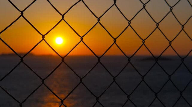 Glowing orange sunset filtering through chain link fence, casting reflective silhouettes across tranquil water surface with golden light symbolizing emotional boundaries and peaceful isolation