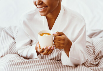 Morning latte, relax and enjoy free time on weekend or self-isolation at covid. Smiling young african american woman sitting on bed with cup of hot coffee, in bedroom interior, copy space, cropped