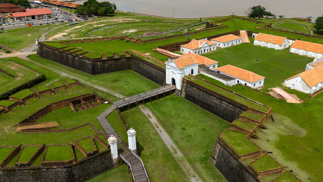 Vista a&eacute;rea de um forte hist&oacute;rico com edif&iacute;cios brancos e telhados alaranjados, cercado por vastas paisagens verdes e um grande rio, exibindo uma mistura de beleza arquitet&ocirc;nica e natural.