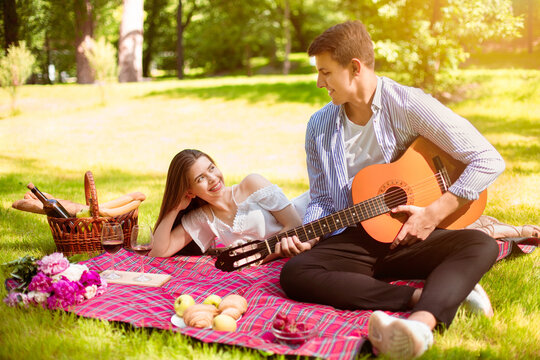 Loving man playing guitar and singing serenade to his girlfriend on romantic picnic at park