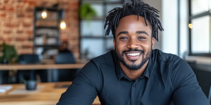 A man with a beard and dreadlocks is smiling at the camera. He is wearing a black shirt and is sitting at a desk