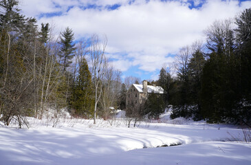 A stone building nestled among bare trees and evergreens in a snowy landscape under a partly cloudy sky with a frozen stream in the foreground