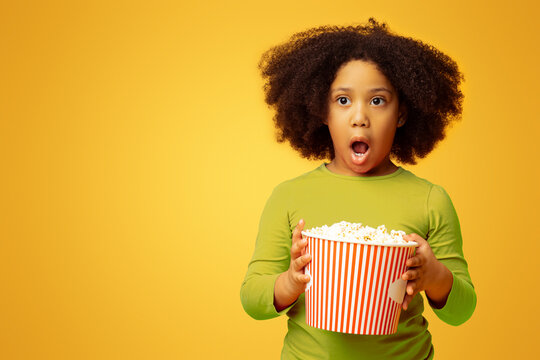 Shocked little african girl eating popcorn from bucket and opening mouth in amazement, standing over yellow background with free space